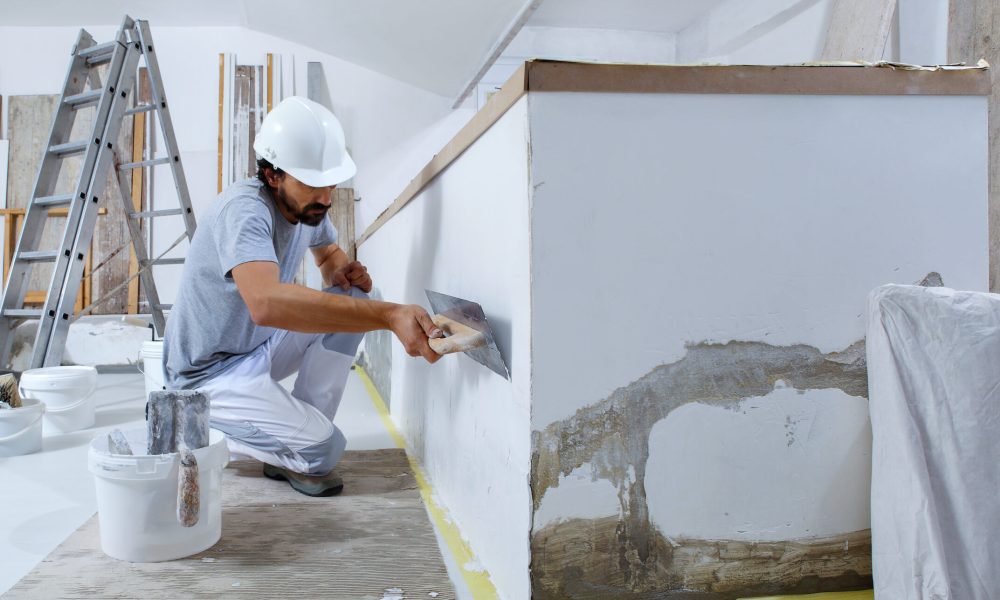 man plasterer construction worker at work, takes plaster from bucket and puts it on trowel to plastering the wall, wears helmet inside the building site of a house